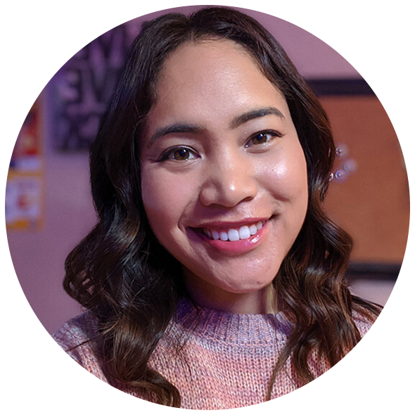 A young woman with shoulder-length wavy dark hair smiling, wearing a pink sweater, in an indoor setting with a purple and orange background.