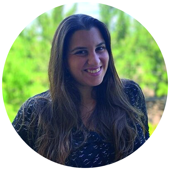 A woman with long brown hair smiling outdoors with a green blurred background.